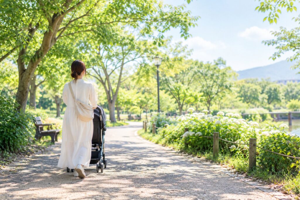 緑の公園の小道を、ベビーカーを押して歩くお母さんの後ろ姿